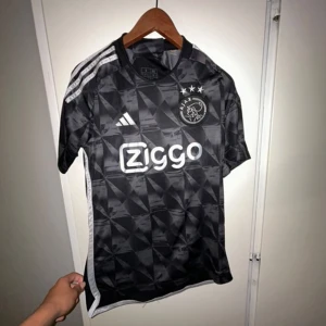 Ajax svart matchtröja Adidas S - Ajax fotbollströja i svart med geometriskt mönster och vita detaljer. Adidas-logga och Ziggo sponsortryck på bröstet samt Ajax klubbmärke med tre stjärnor. Tröjan är kortärmad och tillverkad i ett lätt, tekniskt material som andas.