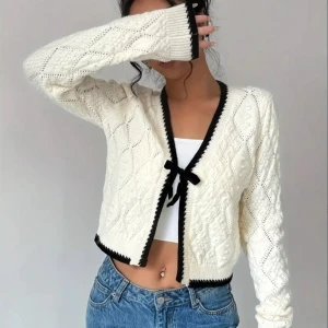 Vit stickad kofta med svart rosett - Supersöt vit stickad kofta med svarta detaljer och rosett i sammet framtill. Koftan är croppad, har lång ärm och ett snyggt mönster i stickningen. Perfekt att slänga över ett linne eller topp för en trendig look. Bara använd 2-3 gånger.