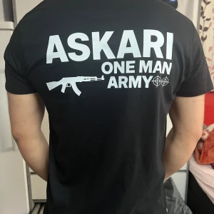 Svart ASKARI t-shirt med tryck - Svart t-shirt med vit text och tryck på både bröstet och ryggen. Stort motiv med texten 'ASKARI ONE MAN ARMY' och en gevärsgrafik på ryggen. Klassisk passform med rund hals och korta ärmar. Perfekt för dig som gillar streetwear och statement-plagg.