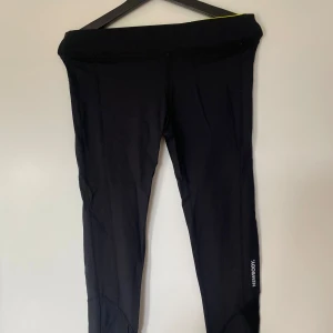 Svarta träningstights från Newbody  - Svarta tights från Newbody med diskret logga på benet och cool meshdetalj vid vaden. Hög midja med insida i neongult. Gjorda i stretchigt material av polyamid och elastan, perfekta för träning eller chill.