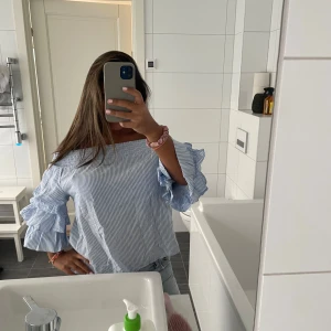 Blåvit randig offshoulder blus - Superfin blus med blåvita ränder och offshoulder-modell. Ärmarna har volangdetaljer och trekvartsärm, vilket ger en trendig och luftig känsla. Perfekt för dig som gillar en söt och somrig stil. Tillverkad i bomull för extra komfort. Storlek 34 men passar även 36 och 38.