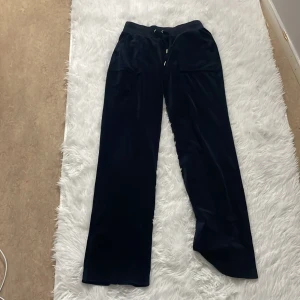 Mörkblå velourbyxor Juicy Couture S - Mörkblå mjukisbyxor från Juicy Couture i glansig velour med broderad logga på ena benet. Byxorna har resår och snörning i midjan, raka ben och fickor både fram och bak med knappdetaljer. Perfekta för en chill och trendig look.