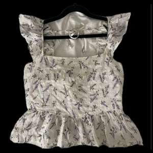 Blommig volangtopp S  - Supersöt topp med volangärmar och blommigt lavendelmönster i lila och grönt på vit botten. Toppen har fyrkantig ringning, smock i ryggen och knytband vid nacken. Perfekt för sommarens alla tillfällen!