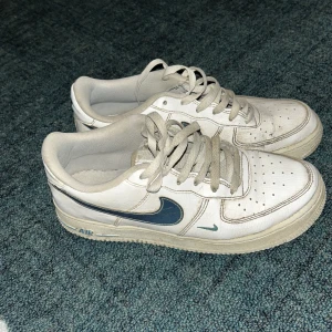 Nike Air Force 1 vita sneakers - Nike Air Force 1 sneakers i vitt skinn med klassisk blå metallic swoosh på sidan. Lågt skaft, perforerad tå och platt sula. Skorna har snörning och diskreta blå detaljer på sula och logga. Perfekt för dig som gillar retrovibbar och streetstyle.