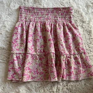Blommig kortkjol från Gina Tricot - Supersöt kortkjol från Gina Tricot med blommigt mönster i rosa, grönt och vitt. Kjolen har smock i midjan (väldigt stretchig) och en volangdetalj. Lite nopprig om man kollar väldigt nära men syns inte annars - tvättas noga inför försäljning! 🩷