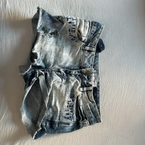 Blå slitna jeansshorts med detaljer - Snygga blå jeansshorts med slitna kanter och fransar. Shortsen har en tvättad look, klassiska fickor och bälteshällor. Dekorativa detaljer med pärlor och paljetter vid fickorna framtill ger extra edge. Perfekta för sommaren och festivalhäng.