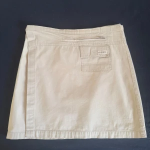 Beige kortkjol från Camel  - Snygg beige kortkjol från Camel. Kjolen har en praktisk ficka med lock och dragkedja framtill samt kardborreknäppning i midjan. Tillverkad i mjuk bomull med enkel, rak passform som ger en chill vibe. Det står storlek XXL men egentligen passar M.