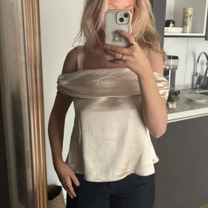 Champagnefärgad omlottopp från NA-KD - Superfin champagnefärgad omlottopp från NA-KD i glansigt satinliknande material. Toppen har breda axelband, offshoulder-detalj och en stor rosett i ryggen. Storlek 34🌸