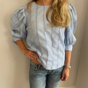 Blårutig blus med puffärm och knyt - Supersöt blus i ljusblått och vitt rutmönster med puffiga trekvartsärmar och knyt vid ärmslut. Avslappnad passform och rund halsringning. Perfekt för en fräsch och trendig look.