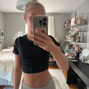 Svart croppad topp med öppen rygg - Svart croppad topp med kort ärm och rund hals. Baksidan har en stor öppen rygg med snörning som knyts längst ner. Toppens material är stretchigt och mjukt, perfekt för en trendig look. Enkel att styla med både jeans och kjol.