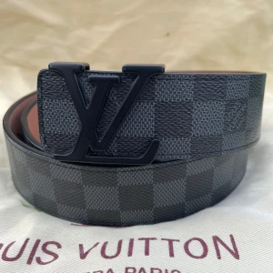 Louis Vuitton Damier Graphite bälte - Snyggt Louis Vuitton bälte i klassiskt Damier Graphite-mönster med svart LV-spänne. Bältet är gjort i exklusivt skinn och har en rutigt design i mörkgrå och svarta toner. Perfekt accessoar för att lyfta din outfit med en lyxig touch.