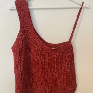 Röd oneshoulder stickad topp från Brownie - Säljer en röd stickad topp från Brownie med glittriga inslag. Toppen har en unik oneshoulder-design med en bred axelrem och en axelbandslös sida.