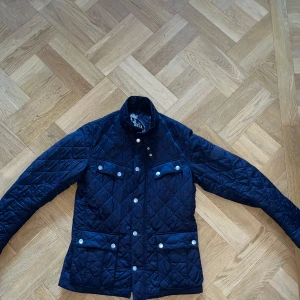Mörkblå quiltad jacka från Barbour - Quiltad mörkblå jacka från Barbour med fyra fickor framtill och coolt motorcykeltryck på insidan. Jackan har tryckknappar, hög krage och är tillverkad i glansig polyester. Perfekt för dig som gillar klassisk men sportig stil.
