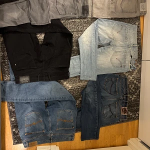 Mix av jeans och byxor i olika färger - Flera par jeans och byxor i olika färger och tvättar, inklusive svarta, gråa, ljusblå och mörkblå. Några har klassiska bakfickor med kontrastsömmar och vissa har slitningar. Perfekt för dig som vill ha variation i garderoben.