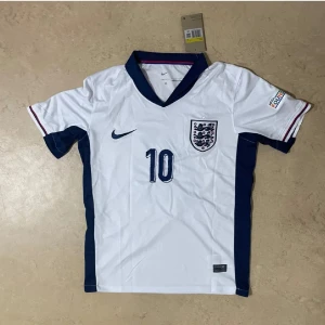 England Bellingham 10 Nike fotbollströja - England fotbollströja med Bellingham och nummer 10. Vit med marinblå detaljer, blå Nike-logga och Englands landslagsmärke på bröstet. Kortärmad modell med blå krage och röda/blå ränder på ärmslut. Tillverkad i lätt och ventilerande material.