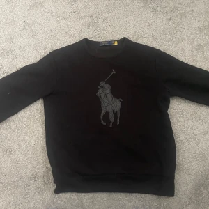 Svart sweatshirt Polo Ralph Lauren - Svart sweatshirt från Polo Ralph Lauren med stort mörkt Big Pony-tryck på bröstet. Klassisk rund halsringning och långa ärmar. Tillverkad i mjuk bomullsfleece som känns riktigt skön och tung. Perfekt för dig som gillar stilren och sportig vibe. Nypris 2799:-