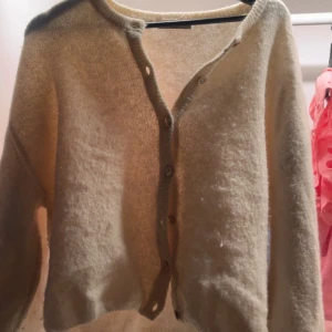 Beige stickad kofta från Gina Tricot - Mysig beige stickad kofta från Gina Tricot i storlek M. Koftan har långa ärmar, rund hals och knappar framtill. Den är croppad och har en mjuk, fluffig känsla som gör den perfekt att slänga på sig över ett linne eller t-shirt.