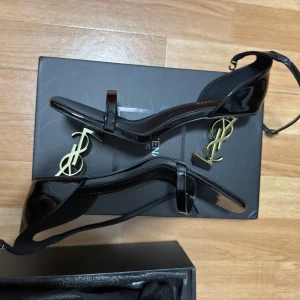 Svarta sandaler med guldklack från Beikenaia - Stilrena svarta sandaler med hög guldklack i metall och öppen tå. Skorna har en smal rem över tårna och runt vristen, samt en fyrkantig tåform. Perfekta för dig som vill sticka ut med en lyxig och modern look.