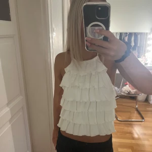 Vit volangtop med knyt i nacken - Supersöt vit topp med flera lager volanger framtill och halterneck med knyt i nacken. Toppen är ärmlös och har en snygg passform som ger en fräsch och somrig vibe. Perfekt för dig som gillar en lekfull och trendig stil. Använd 1 gång 