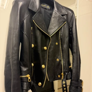 Balmain x H&M svart skinnjacka - Säljer en svart skinnjacka från Balmain x H&M med guldiga knappar, dragkedjor och bältesdetaljer. Jackan har klassisk bikerstil med bred krage, dubbla rader knappar och justerbara spännen vid ärmslut och midja. Perfekt statement-plagg för dig som gillar edgy mode.