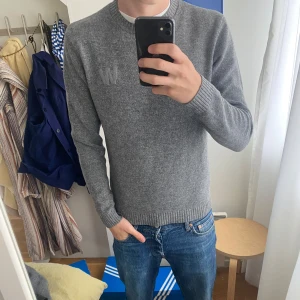 Grå stickad tröja från Wood Wood - Säljer en stilren grå stickad tröja från Wood Wood med broderad logga på bröstet. Tröjan har rund halsringning, ribbade muddar och är långärmad. Perfekt för lager-på-lager och funkar året runt.