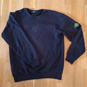 Svart sweatshirt från Stone Island - Snygg svart sweatshirt från Stone Island med klassisk rund hals och broderad logga på bröstet. Den ser ut att vara mörkblå på bilden men den är svart. Ikonisk patch på vänster ärm. Mjuk bomullskvalitet och ribbade muddar vid ärmslut och nederkant. Perfekt för en clean och stilren look.