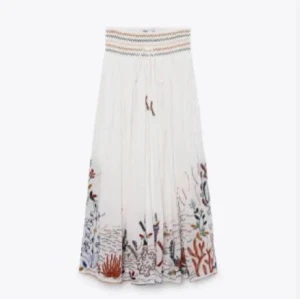 Vit långkjol med broderi från Zara - Supersöt vit långkjol från Zara med broderade detaljer längst ner i färgglada nyanser. Kjolen har bred resår i midjan med vågigt mönster och snörning framtill. Perfekt flowy och luftig för varma dagar.