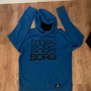 Blå hoodie från Björn Borg - Snygg blå hoodie från Björn Borg med stor svart logga på bröstet. Tröjan har huva, lång ärm och en stor magficka framtill. Perfekt för chill eller träning, och tillverkad i mjuk bomullsmix som känns skön mot huden.