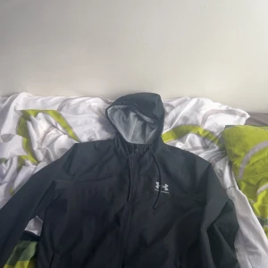 Svart vindjacka från Under Armour - Svart vindjacka från Under Armour med huva och dragsko. Jackan har dragkedja framtill, meshfoder och logga på bröstet. Perfekt för blåsiga dagar och sportiga outfits. Snygg, enkel och funktionell design som passar till allt.