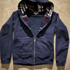Marinblå hoodie från Burberry - Snygg marinblå hoodie från Burberry med klassiskt rutigt foder i huvan och längs dragkedjan. Tröjan har huva, dragkedja framtill och två stora fickor. Perfekt för dig som gillar stilrena och exklusiva streetwear-plagg. Hör av vid minsta fundering 💫