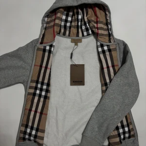 Grå Burburry zip hoodie  - Snygg grå hoodie från Burberry med klassiskt rutigt foder i huvan och insida. Tröjan har dragkedja framtill, snörning vid huvan och broderad logga på bröstet. Perfekt för dig som gillar stilrena och exklusiva plagg med en twist.