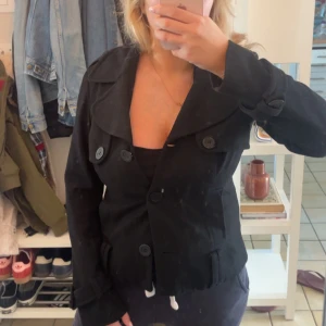 Svart kappa - Snygg svart kappa med stora knappar och bred krage. Jackan är kortare i modellen och har långa ärmar med knappdetaljer vid handlederna. Perfekt för dig som gillar en stilren och enkel look med en twist.