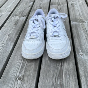 Nike Air Force 1 vita sneakers - Klassiska Nike Air Force 1 sneakers i helvitt skinn med perforerad tå, rund tå och snörning. Snygg och tidlös modell med låg profil och ikonisk swoosh på sidan. Perfekta för dig som gillar streetstyle och vill ha en clean look.