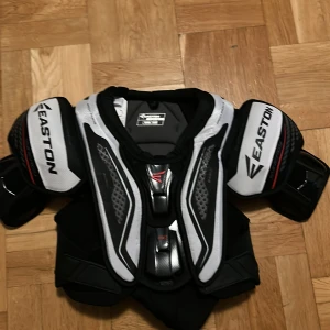 Easton Synergy hockey axelskydd M - Säljer en Easton Synergy hockeyväst i storlek M/Senior. Västen är svart och vit med coola grafiska detaljer och extra skydd vid axlar och bröst. Perfekt för dig som lirar hockey och vill ha bra skydd och grym passform.