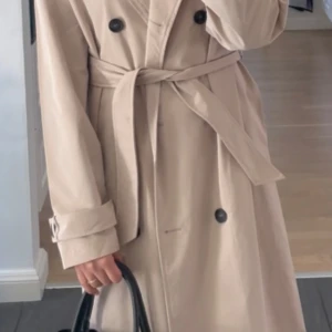 Beige trenchcoat med bälte och knappar - Stilren beige trenchcoat med dubbelknäppning och bälte i midjan. Jackan har långa ärmar, klassisk krage och svarta knappar som kontrast. Perfekt för dig som vill ha en tidlös och snygg look till vardags. Passar perfekt som höst/sommar kappa. Lätt att matcha med olika outfits. Använd endast 1 gång. Köptes från Zalando.