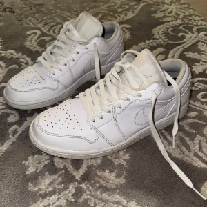 Nike Air Jordan 1 Low  - Snygga Nike Air Jordan 1 Low i helvitt skinn med klassisk siluett och perforerad tå. Skorna har vit sula, vita skosnören och diskret broderad logga på hälen. Perfekta för dig som gillar stilrena och tidlösa sneakers.