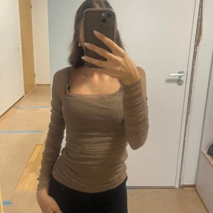 Brun glittrig långärmad topp - Säljer en brun glittrig topp med långärmade, åtsittande ärmar och rynkade detaljer längs sidorna. Toppen har fyrkantig ringning och är croppad i modellen. Perfekt för dig som vill sticka ut lite extra med en snygg shimmer-effekt.