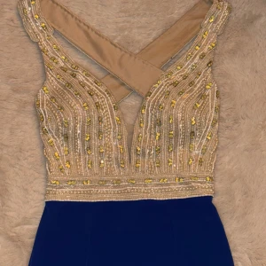 Blå långklänning med glittrig topp - Säljer en elegant långklänning med djup v-ringad och korsad rygg. Toppen är champagnefärgad med glittriga pärlor och paljetter i guld och silver, medan kjolen är marinblå och slät. Perfekt för bal eller fest! Klänningen har axelbandslös design och smickrande passform.