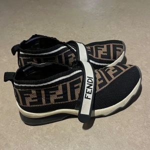Fendi sneakers med FF-mönster strl 37 - Säljer ett par snygga äkta Fendi sneakers i svart och brunt med det klassiska FF-mönstret. Skorna har en vit och svart rem med texten 'FENDI' och 'LOVE'. Skorna kommer i super bra skick! Låda medföljer. 