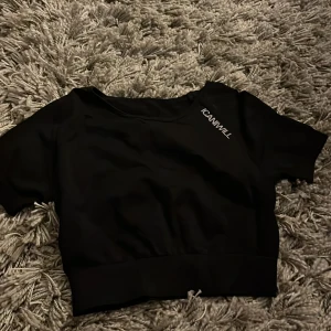 Svart croppad topp från ICANIWILL - Svart croppad topp från ICANIWILL med korta ärmar och bred resår nedtill. Toppen har en stilren design med logga tryckt på bröstet och är gjord i ett stretchigt syntetmaterial som passar perfekt till träning.