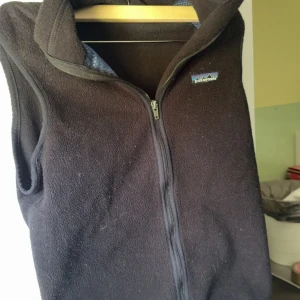 Svart fleeceväst från Patagonia - Svart fleeceväst från Patagonia. Västen har en liten logga på bröstet och en praktisk ficka med dragkedja på ryggen. Perfekt för lager-på-lager och chill vardagsstil.