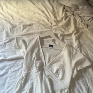 Vit omlottklänning från Gina Tricot - Supersöt vit kortklänning från Gina Tricot med omlott-design och knyt i midjan. Klänningen har korta ärmar och v-ringad hals, perfekt för sommarens alla tillfällen. Tillverkad i mjuk bomull som känns skön mot huden.