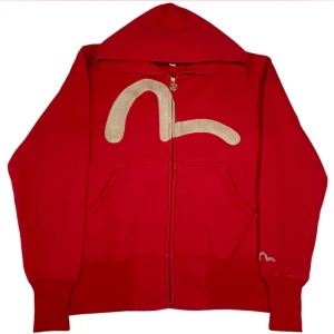 Sällsynt evisu zip up  - Säljer en röd hoodie från Evisu med stor beige broderad detalj på framsidan och mindre broderi på ärmen. Tröjan har huva, dragkedja och fickor framtill. Unik design med coolt myntformat dragkedjesmycke. Perfekt för dig som gillar streetwear.