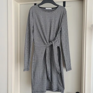 Svartvit rutig klänning Mango S - Snygg svartvit klänning från Mango i storlek S med klassiskt rutigt mönster. Modellen är kort med lång ärm och har en cool knytning i midjan som ger extra stilpoäng. Perfekt för dig som gillar tidlös design med en twist.