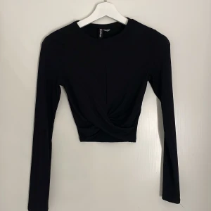 Svart croppad topp från H&M Divided - Svart långärmad croppad topp från H&M Divided i ribbat material. Toppen har en snygg twistad detalj framtill som ger en cool look. Perfekt till en trendig outfit.