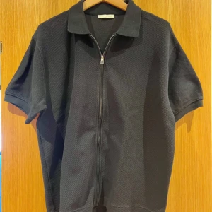 Svart zip-tshirt med krage från Zara - Svart t-shirt från Zara med korta ärmar och cool krage. Framsidan har en dragkedja hela vägen och materialet är strukturerat och känns som bomull. Perfekt för dig som gillar stilrena och enkla plagg med en twist.