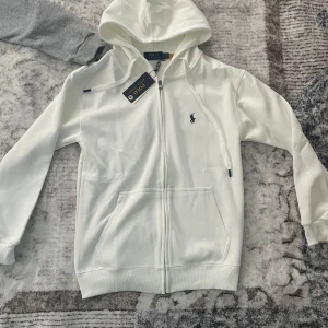 Vit hoodie från Polo Ralph Lauren - Snygg vit hoodie från Polo Ralph Lauren med dragkedja och huva. Klassisk logga broderad på bröstet och stora fickor framtill. Tillverkad i mjuk bomullsmix, perfekt för en chill och stilren look.