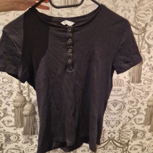 Svart ribbad topp med knappar - En svart ribbad topp med korta ärmar och rund hals. Toppen har fyra dekorativa knappar framtill som ger en cool detalj. Perfekt till jeans eller kjol för en avslappnad stil.