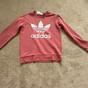 Rosa Adidas sweatshirt med logga - Snygg rosa sweatshirt från Adidas med stor vit trefoil-logga och text framtill. Tröjan har rund hals, långa ärmar och är gjord i mjuk bomullsmix. Perfekt för en sportig och avslappnad stil.