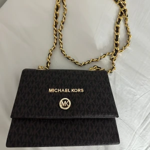 Svart axelväska från Michael Kors - Snygg svart axelväska från Michael Kors med guldiga kedjedetaljer och logomönster över hela väskan. Väskan har ett lock med magnetlås och rymligt fack inuti. Perfekt för dig som vill ha en stilren och trendig accessoar. Aldrig använt 
